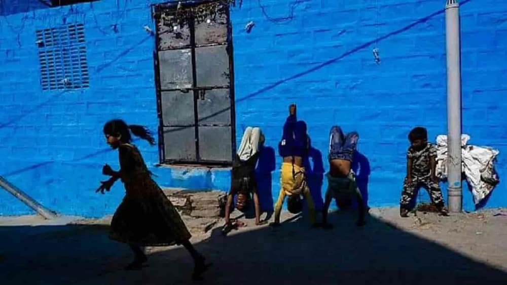A photo shows some children having fun on the side of a street. While some are doing handstands against the blue wall, a younger boy watches, and a girl in the foreground runs away laughing. This is one of the photos in the street photography exhibition at the International Center of Photography.