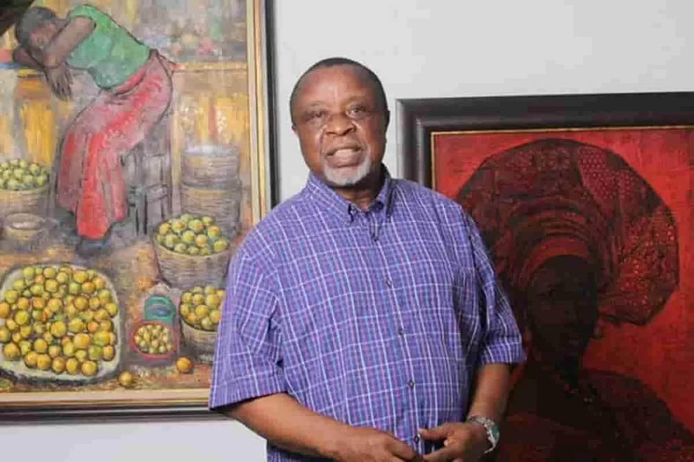 Kolade Oshinowo Nigerian artist wearing a blue shirt, stands in front of two of his colorful paintings, smiling.