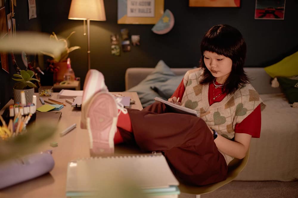 A woman sitting in her bedroom with her feet up on her desk full of crafting supplies. She is writing in a notebook.