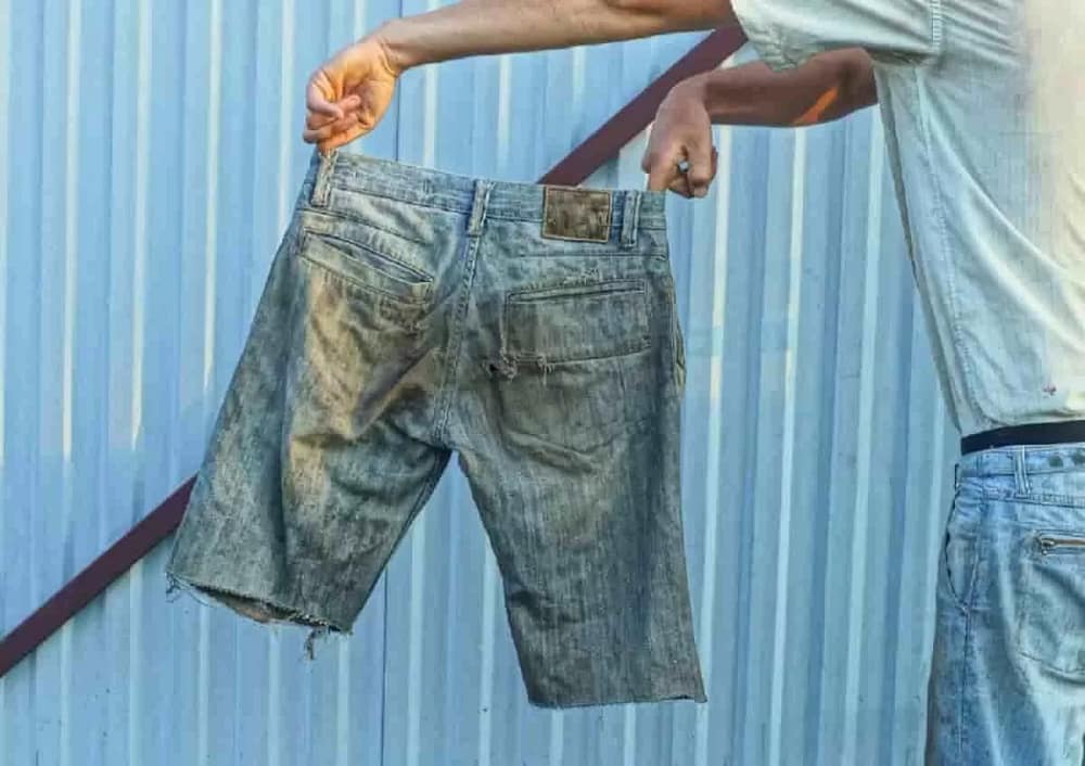 Someone holding a pair of distressed and deconstructed denim shorts off to their left side. The shorts are ripped and dirty.