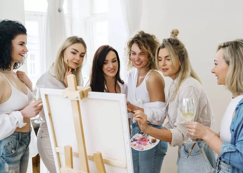 A group of women, with glasses of wine in hand, watch their friend showcase her artistic skills at an at-home art party.