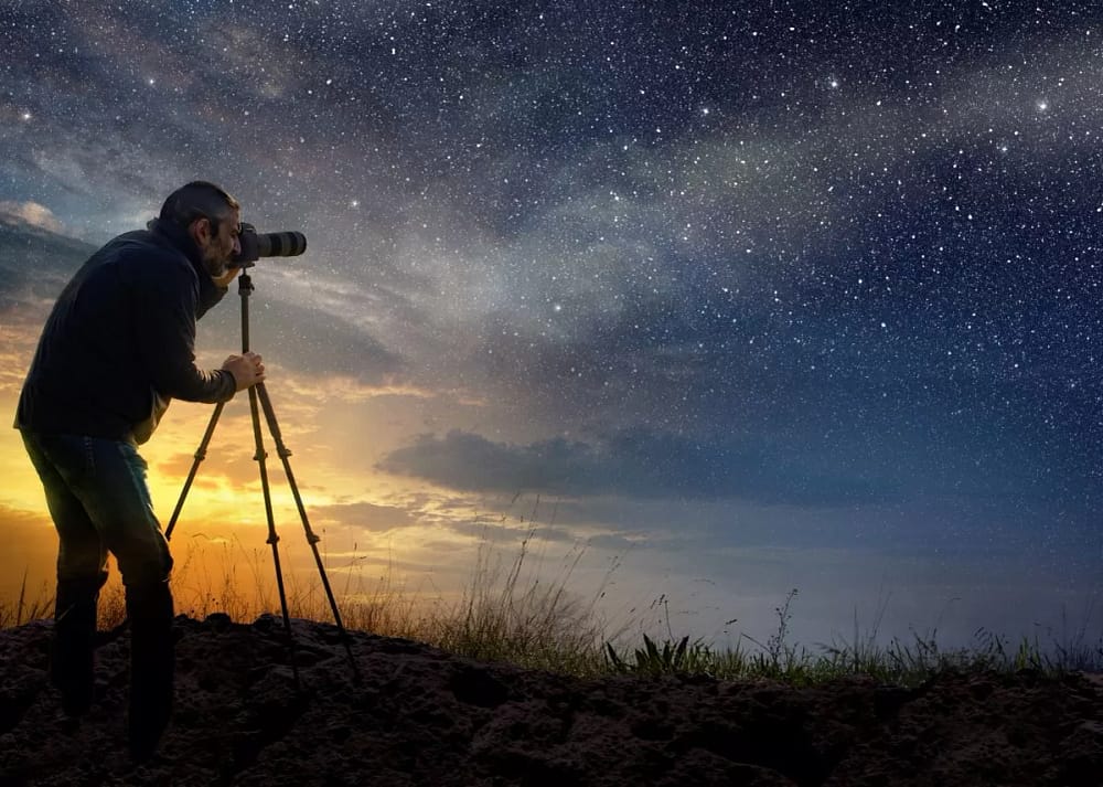 A man uses a camera on a tripod to take pictures of the sky with the faint sun on one side and the starry sky ahead. Night photography requires patience