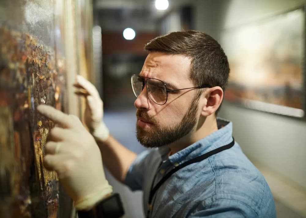 A bearded man in a denim shirt, white protective gloves, and glasses inspects a painting he plans to restore. Restoring painting is one of the artist side hustles that pay well and is super fun.