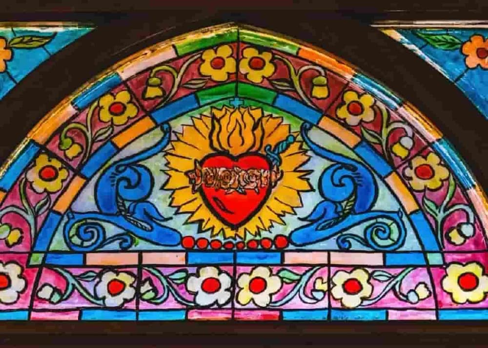 Multicolored stained glass features the Sacred Heart at its center. The heart is wreathed with roses and surrounded by flames. Common key symbols in Catholic art have significant meanings