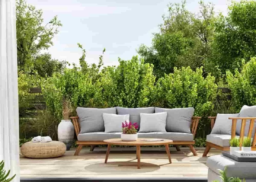 The sliding doors of a home are open. Light shines on a patio area with wooden furniture, gray cushions, and large shrubs. You can transform your-patio with expert exterior-design tips