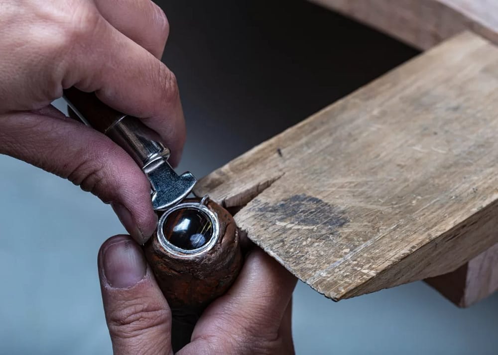 Close-up of two hands holding lapidary tools with a round brown crystal rock next to a wooden base with a small opening. Enhancing gemstone beauty is one of the reasons why precise rock cutting is crucial in lapidary.