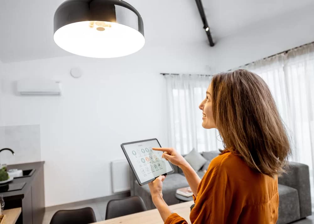 A woman stands in the home kitchen, touching the tablet screen and looking at a light hanging from the ceiling.