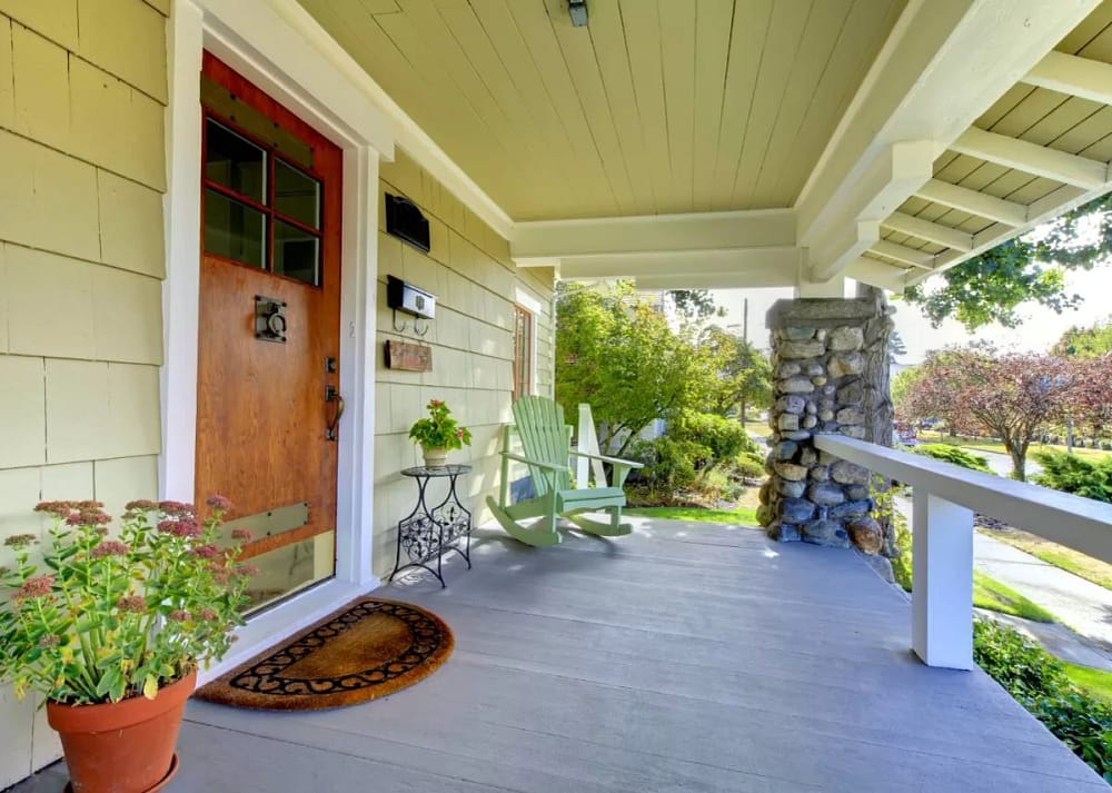 A covered front porch contains a large potted plant, a green rocking chair, and a small black table with a small potted plant.