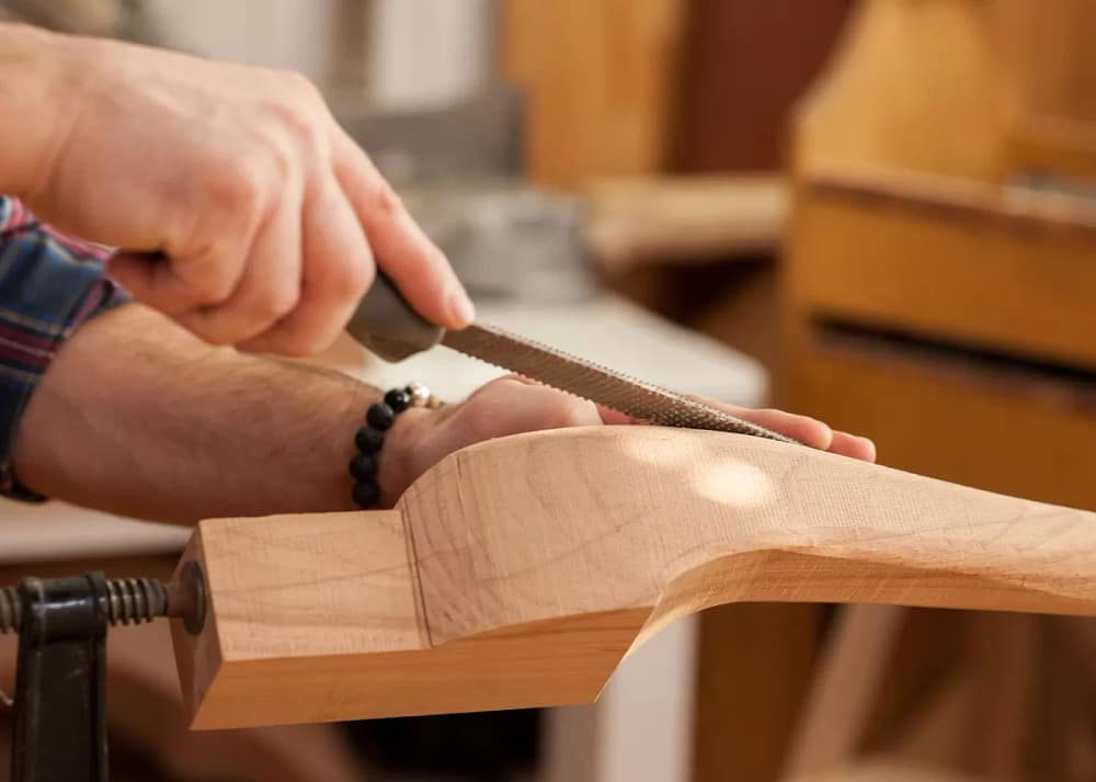A close-up of a man's hands using a metal tool to work on a piece of wooden furniture. There are other wooden pieces in the background.