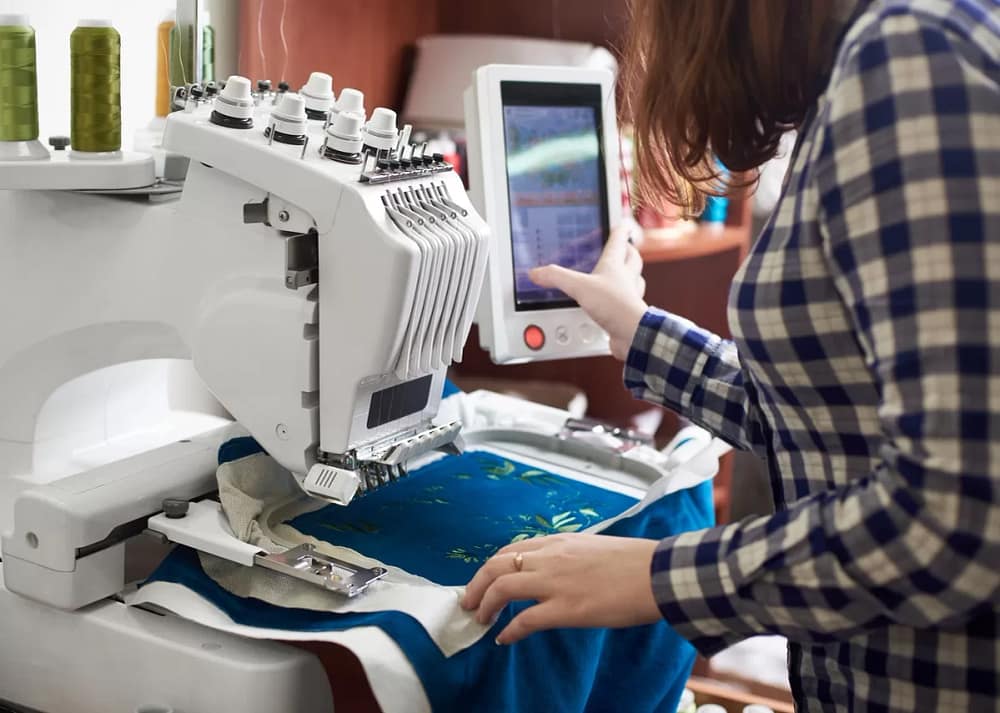An embroidery business owner standing at the machine programming it to embroider a blue garment placed in a hoop.
