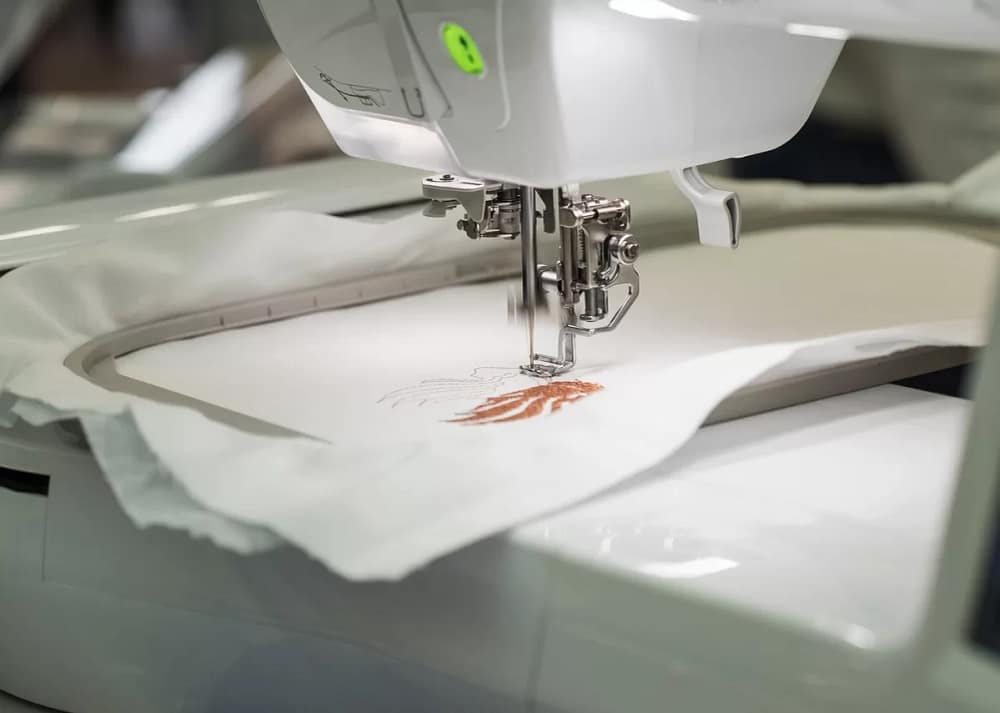 An embroidery machine embroidering a detailed pattern onto a piece of white fabric using copper-brown thread.