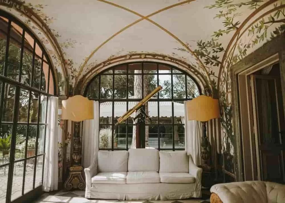 A classic Renaissance-style home design with a sofa, wide windows, and a telescope. There's gold detailing in the ceiling. An important example of creating harmony with contrasting design elements