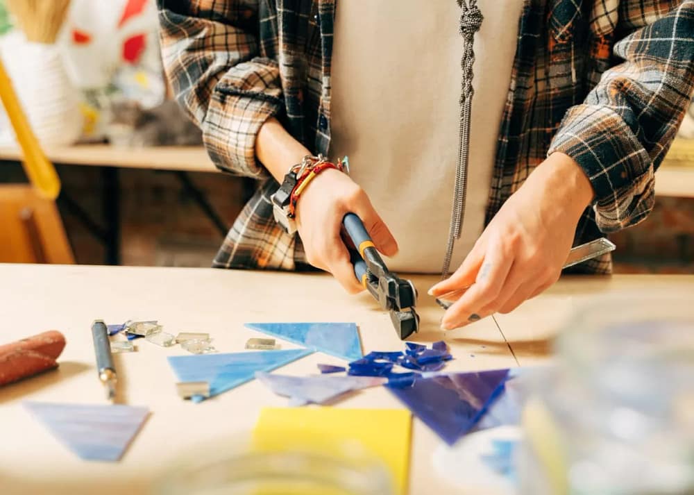 A close-up of a woman is in her art studio, putting together a glass mosaic with tweezers and a fusing tool.