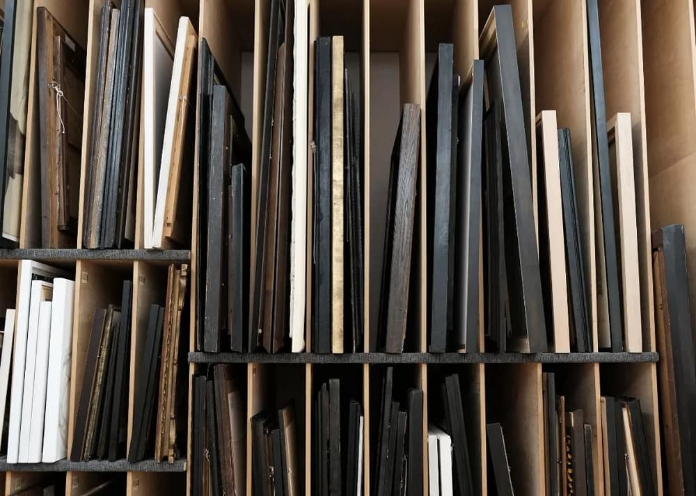 Storage shelves with assorted framed artworks and canvases organized vertically in narrow wooden rack compartments. A good gallery storage and organization will protect your art.