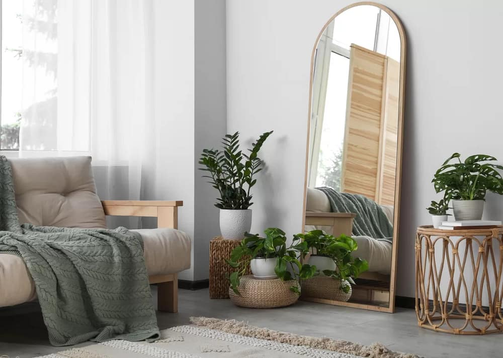 A living room with a tan futon and green blanket. Several plants rest on baskets, with a tall mirror resting against a wall. Plants and mirrors are design choices that make homes feel more spacious