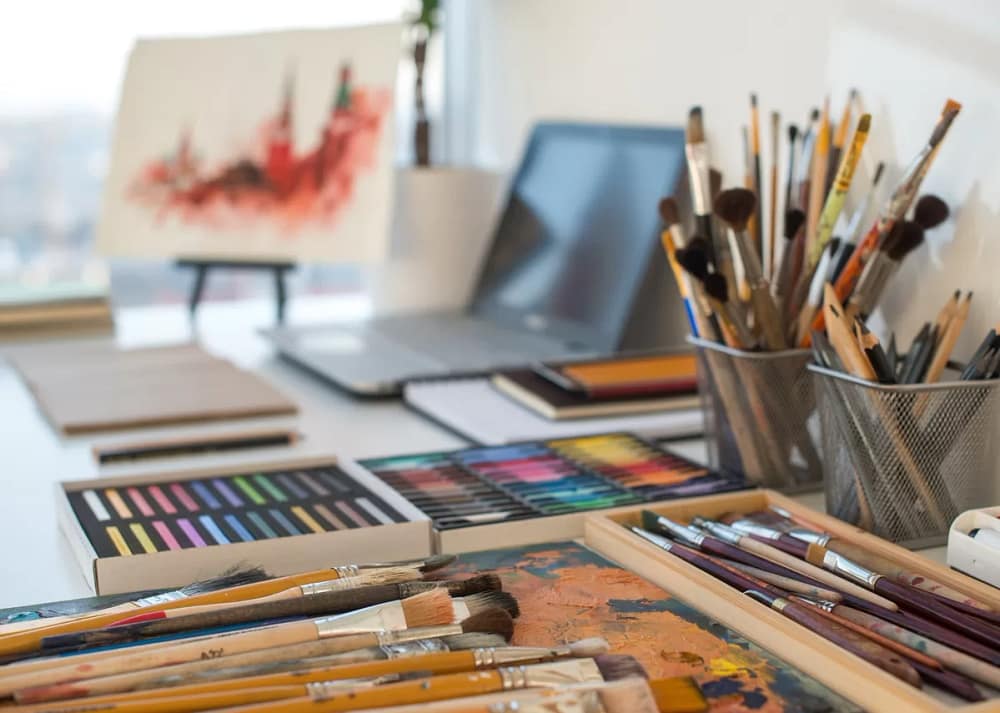 A white table is filled with art materials like paintbrushes, pencils, crayons, and an easel with a painting. Use practical ways to create more creative space at home
