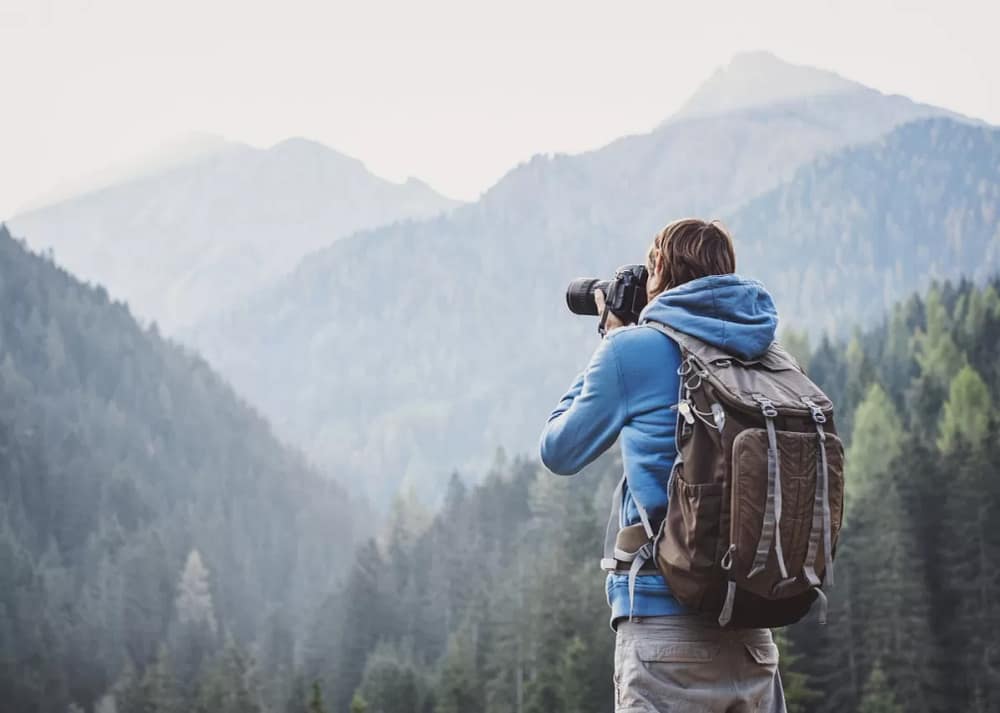 A young photographer stands with his camera facing outwards toward a large expanse of trees in the distance. One of the tips for mastering composition in your photos is to avoid standing still