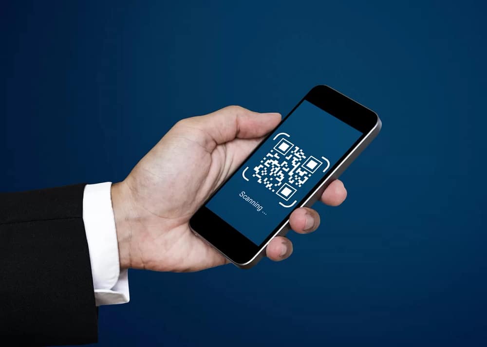 A hand in formal wear holds a smartphone displaying a QR code with text stating "Scanning..." on a dark blue background. Engaging visitors with QR codes on museum signage is an effective way to captivate audiences.