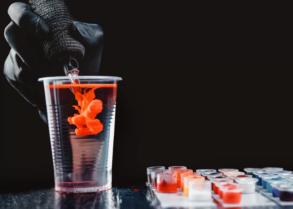 A gloved hand lowering a wrapped rotary pen tattoo machine into a cup of clear water to wash orange ink out of the needles.