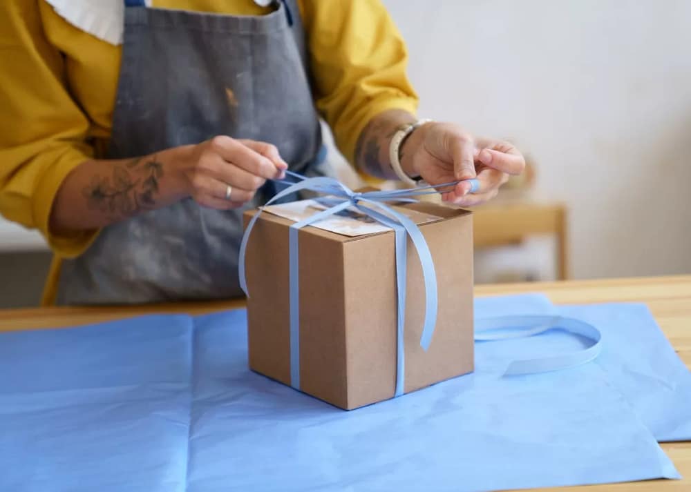 A person wearing a dirty crafting apron while tying a thin blue ribbon around a cardboard gift box on a table. A gift guide for creatives will help you select the best give for that painter