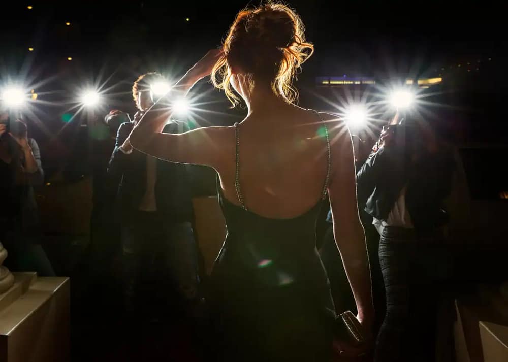 A rear view of a woman with a high bun shields her face from a crowd of people holding lights and cameras.