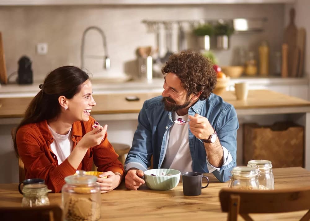 Two people sitting down at a table in a welcoming residential kitchen. Both people smile while eating bowls of fruit.