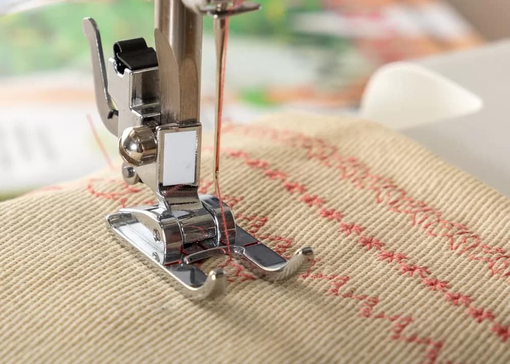 A close-up of a needle stitching zig-zagged rows in red thread on tan fabric through the sewing machine's presser foot. This just one of the types of sewing machine feet and their uses