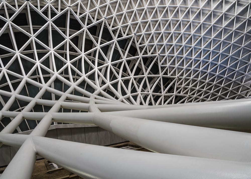 A white geometric lattice facade with interwoven metal beams forming a complex pattern over a glass structure.