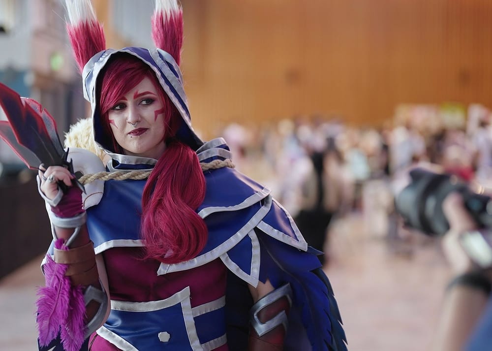 A woman in a blue cape and hood poses for a photo in a large convention hall with a crowd visible in the background.