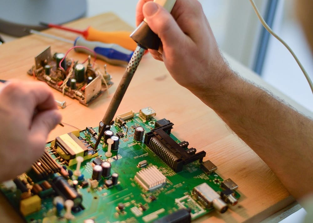 A person repairs a circuit board with a soldering iron while preparing old electronics for a mixed-media art project. This preparation is an important step in how to build mixed-media art with old electronics.