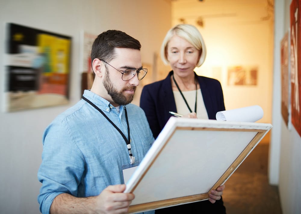 A bearded man holds a framed canvas while a woman writes on a clipboard in an art gallery hallway, illustrating the important questions to ask before buying fine artwork.