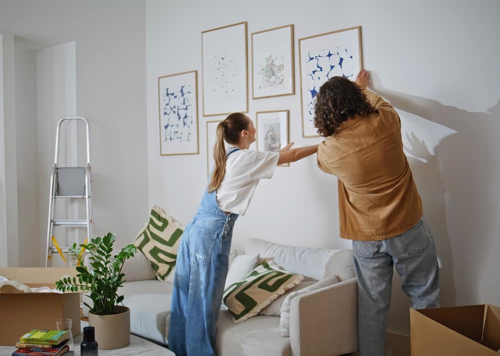 a man and woman standing by a white wall while hanging framed pictures above a sofa in a bright living room.