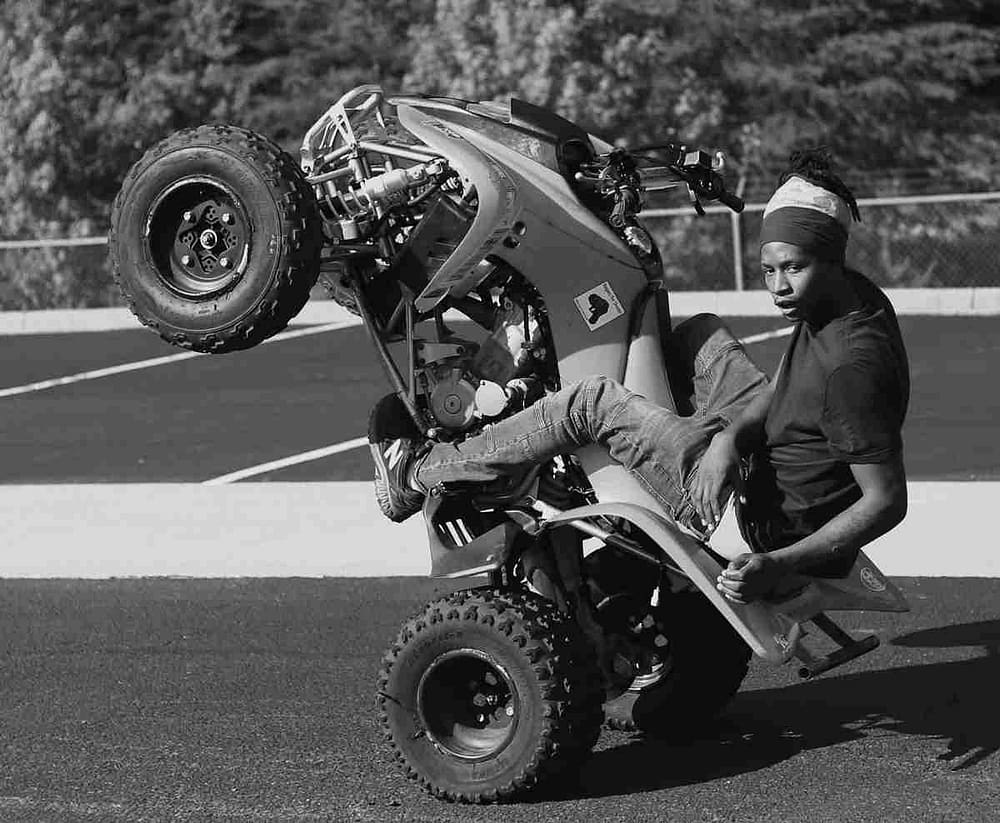 Image: Two Wheels 2 God, a 2019 black and white photograph of a young man on an ATV popping wheelie, in Godspeed an intimate encounter of DC BikeLife