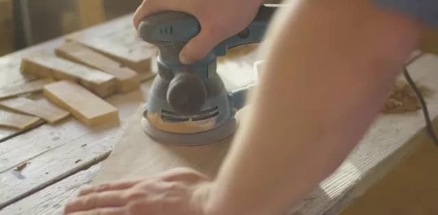 The hands of a woodworker are using an orbital sander to sand down the surface of a piece of wood he's working on.