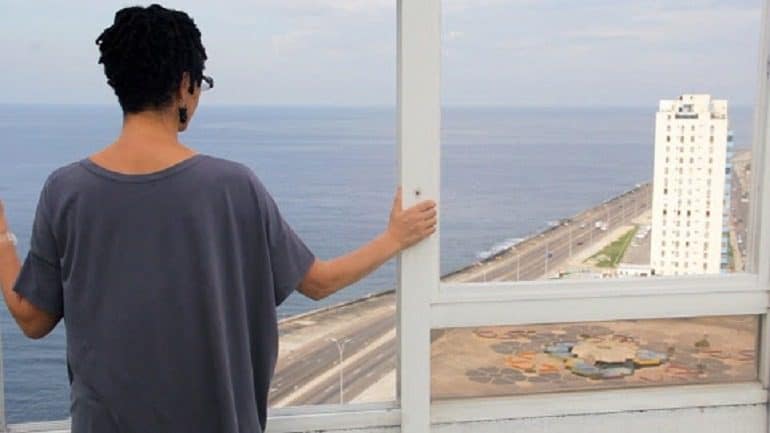 Image: A woman stands at the window of a skyscraper looking at tall buildings in a Still from Coco Fusco, La Confesión, a 30-minute single channel digital film made in 2015