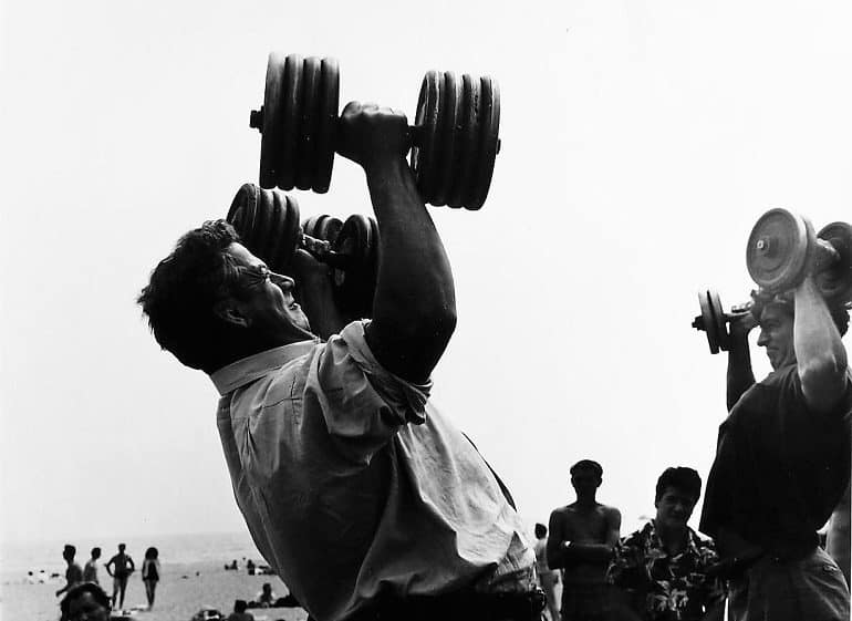 Image: A black and white photograph of a Man with Dumb-bells at the Muscle Beach, Santa Monica, CA, 1954 by Larry Silver