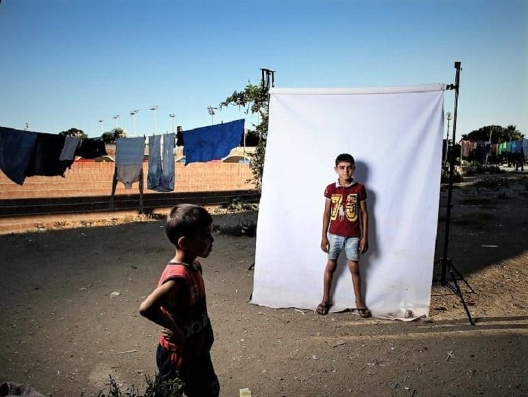 Image: Photograph of a boy wearing a red polo tee shirt with number 75 boldly written on it in front of a white background is one of the photographs by Iranian photographer Amir Kabir Jabbari highlighting the lives of Syrian Civil War children